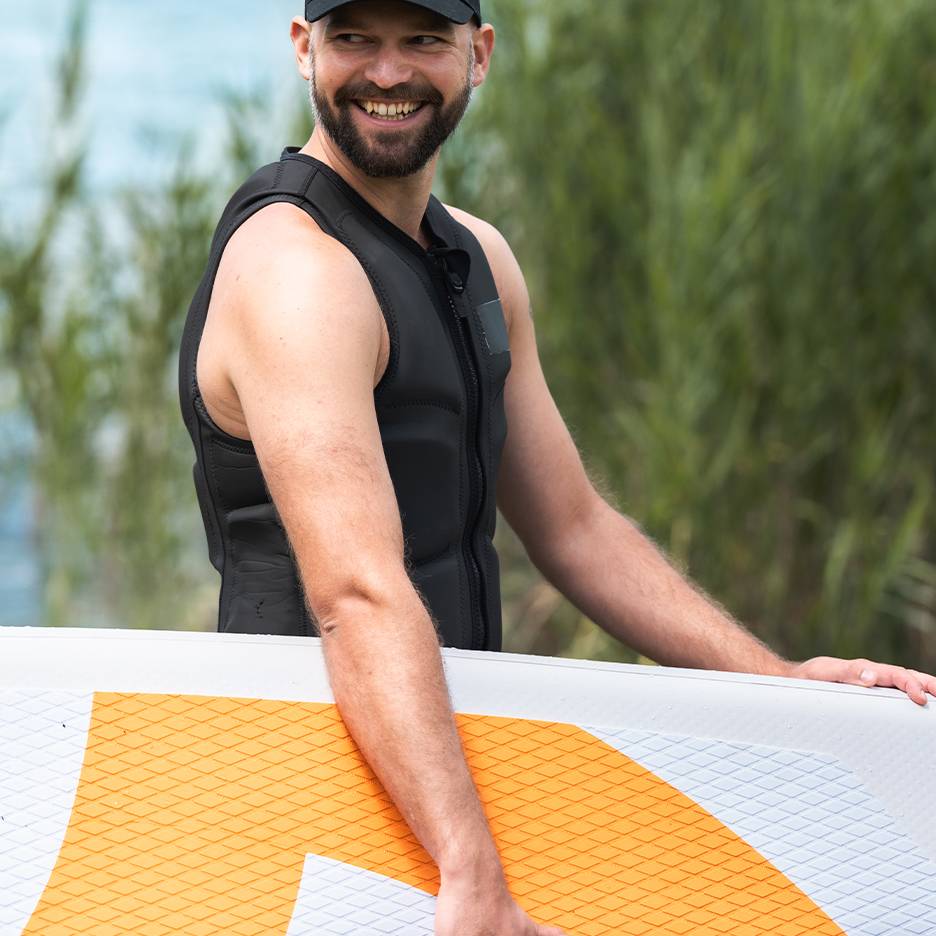Homme souriant en gilet de sauvetage noir tenant une planche de paddle.