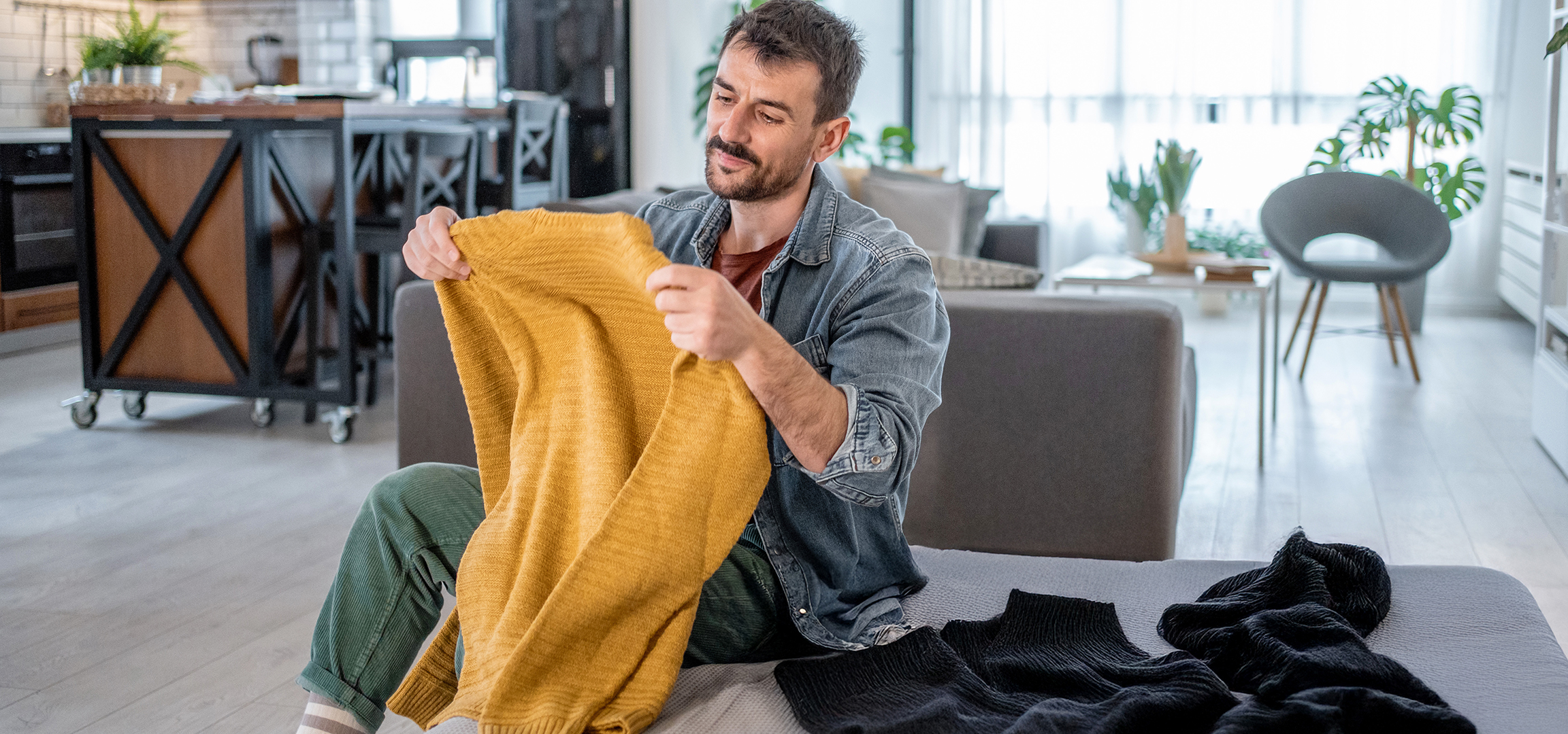 Homme tenant un pull en tricot jaune, avec d'autres pulls sur un canapé dans un salon.