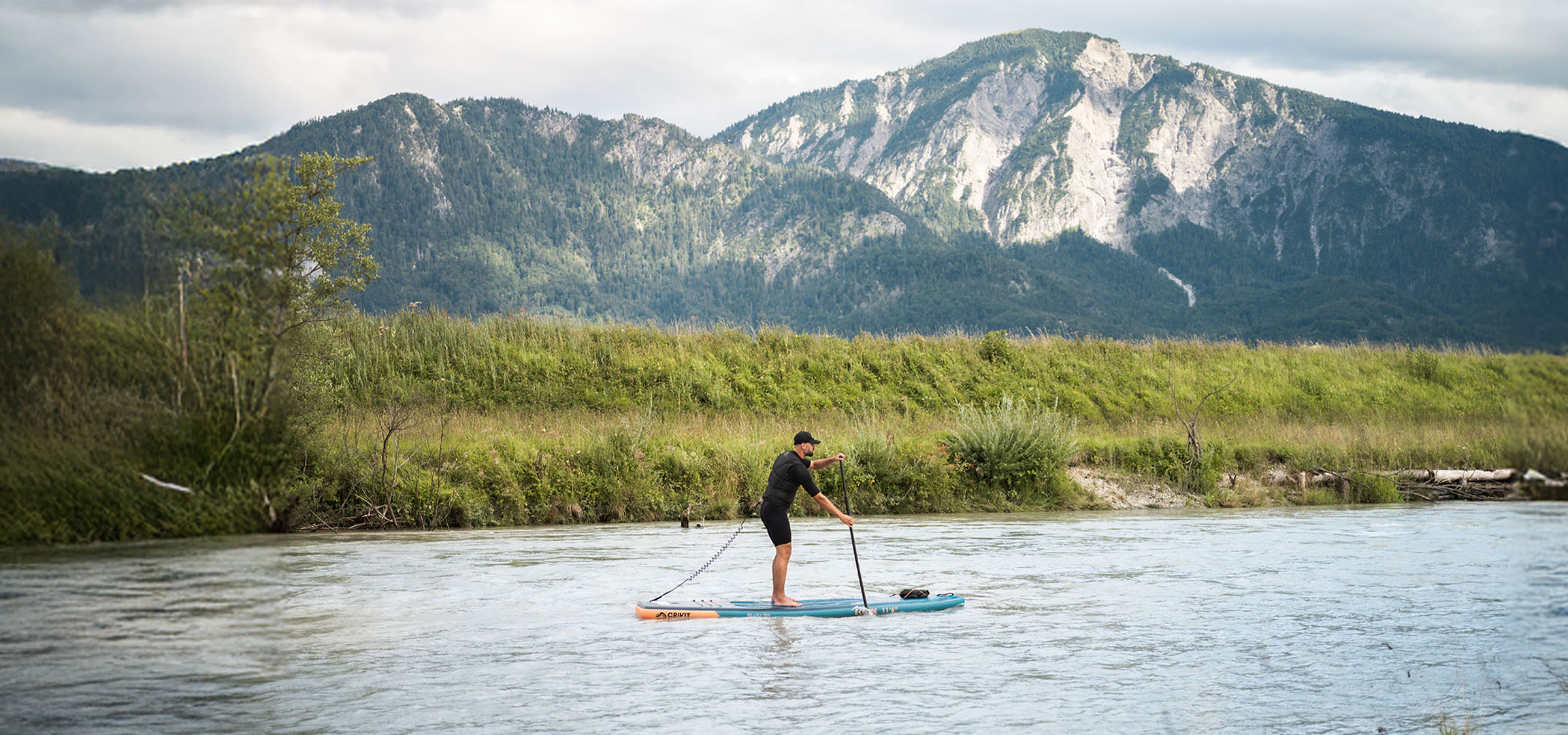 Homme en paddleboard sur une rivière avec des montagnes et une végétation luxuriante en arrière-plan.