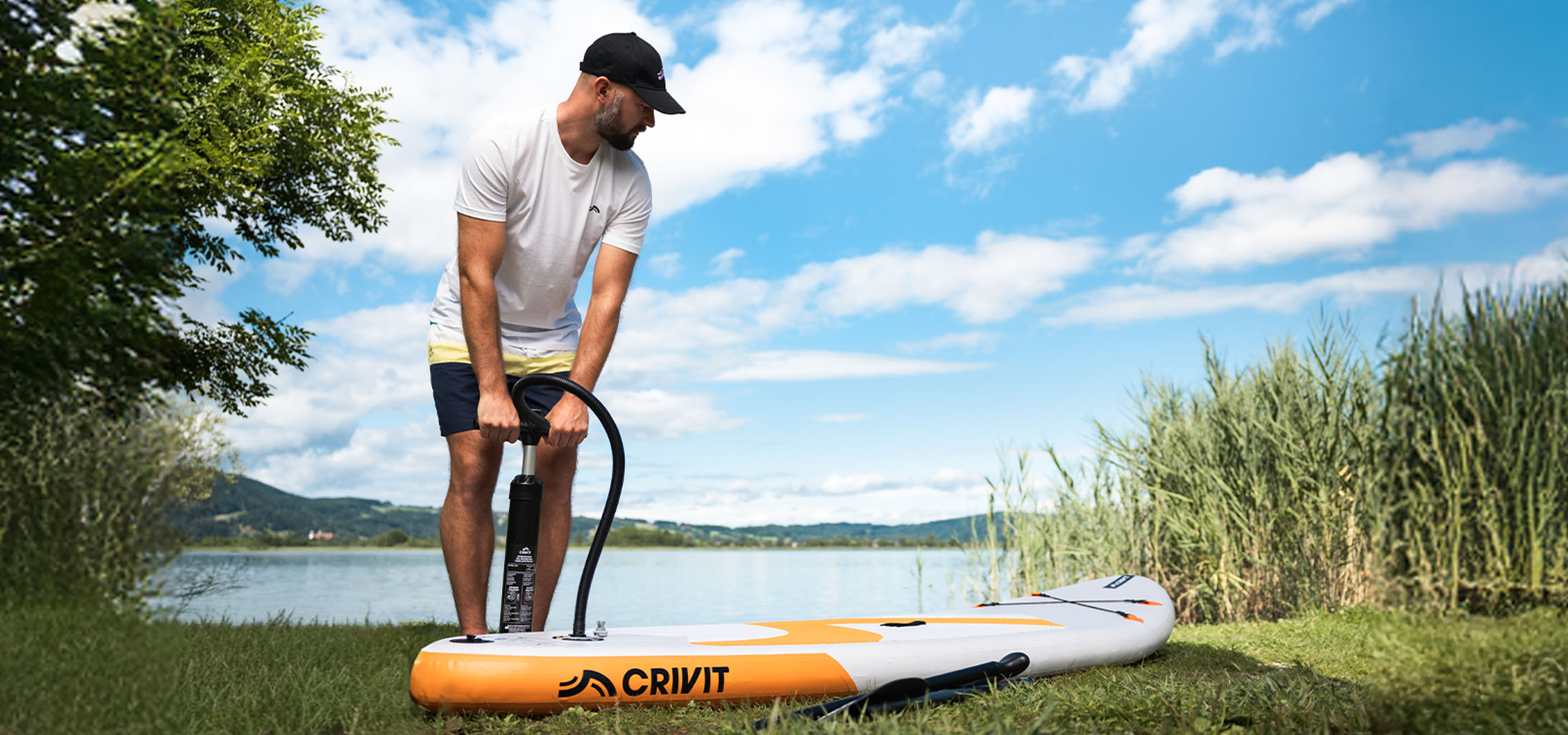 Homme gonflant une planche de SUP CRIVIT au bord d'un lac sur l'herbe verte.