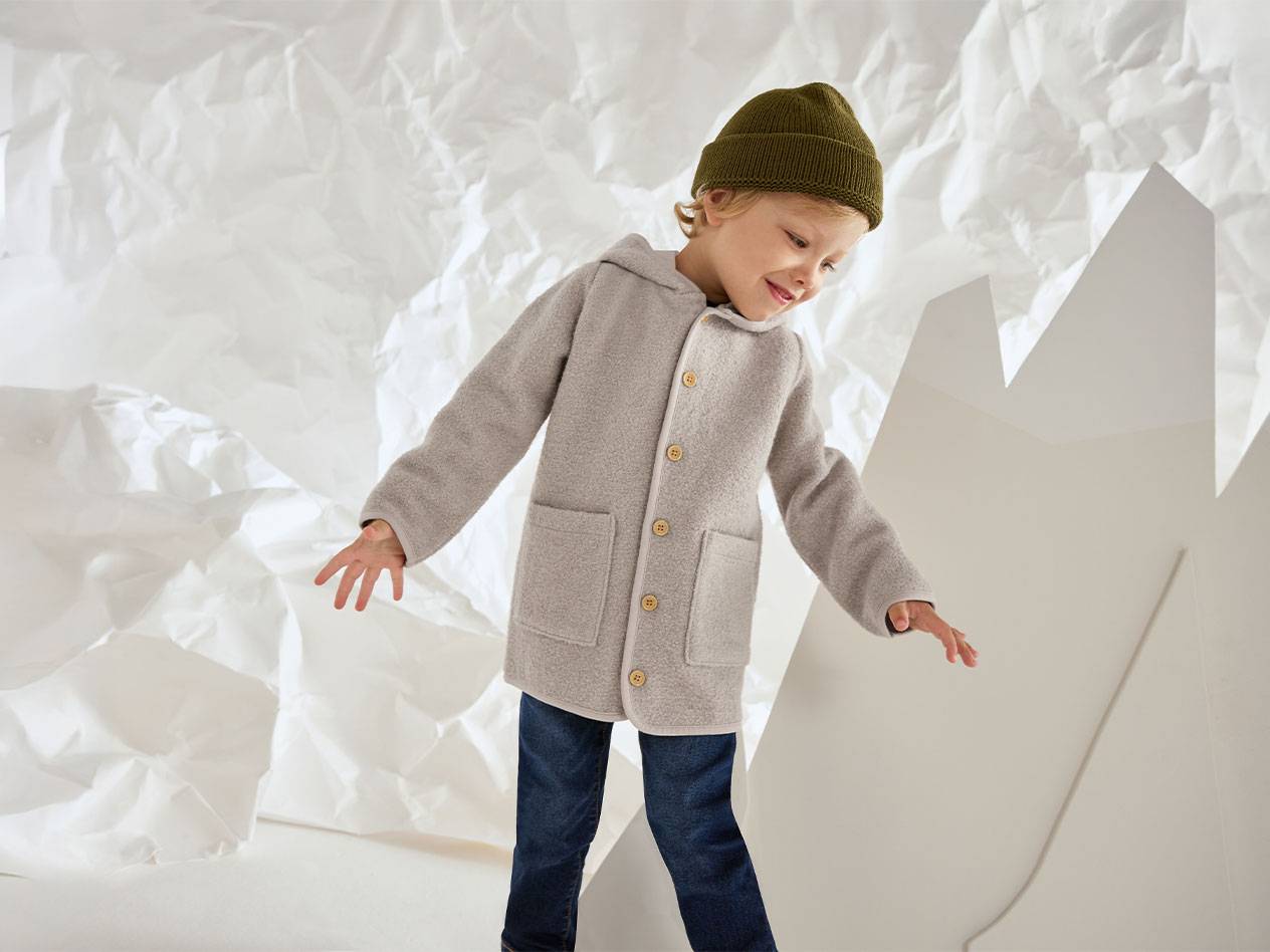 Garçon en veste de laine grise et bonnet vert, souriant sur un fond de papier froissé.