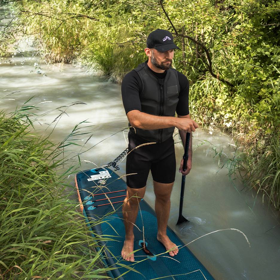 Homme sur une planche de paddle dans une rivière, portant une combinaison et un gilet de sauvetage.
