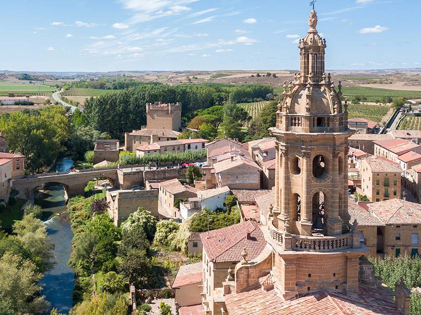 Vue aérienne d'une ville historique avec un clocher proéminent, une rivière, un pont et des vignobles.
