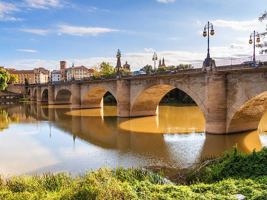 Pont en arc de pierre sur une rivière avec une ville et un ciel bleu en arrière-plan.