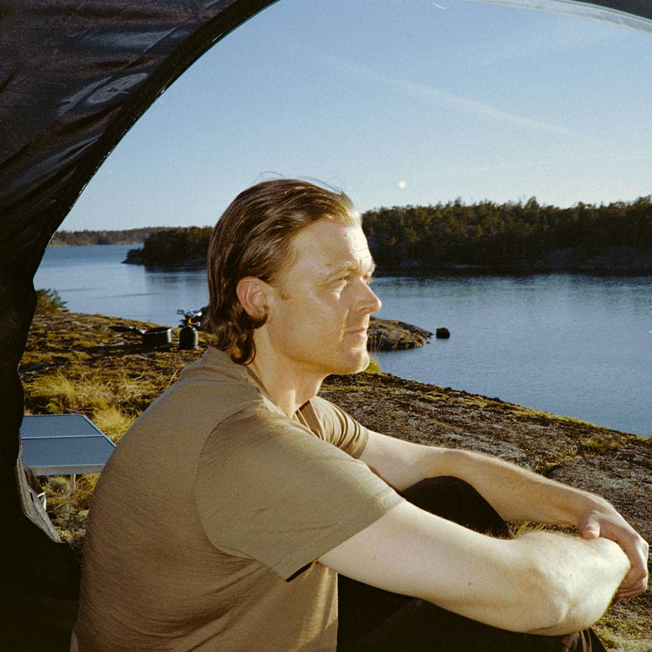 Homme en t-shirt de sport assis dans une tente, regardant un lac et une forêt.