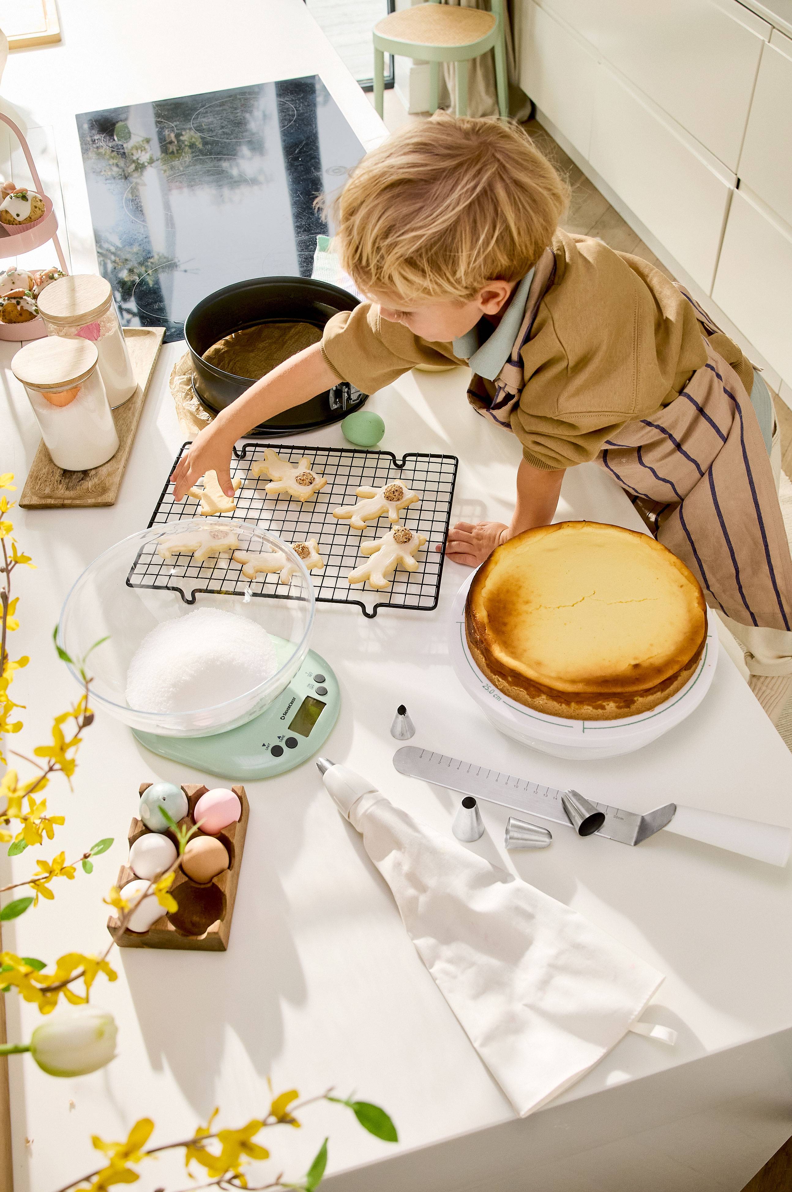 Un enfant décore des biscuits de Pâques, avec un cheesecake et des ustensiles de pâtisserie sur la table.