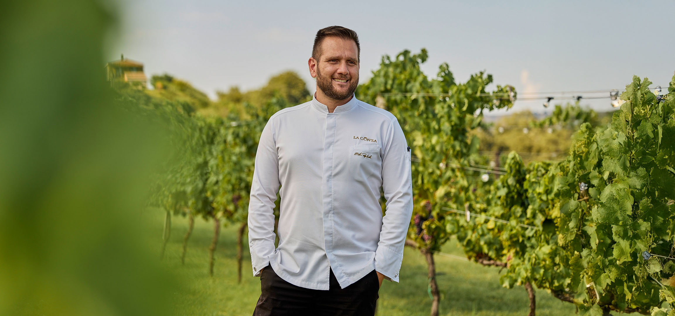 Un homme souriant en veste de chef blanche dans un vignoble ensoleillé.