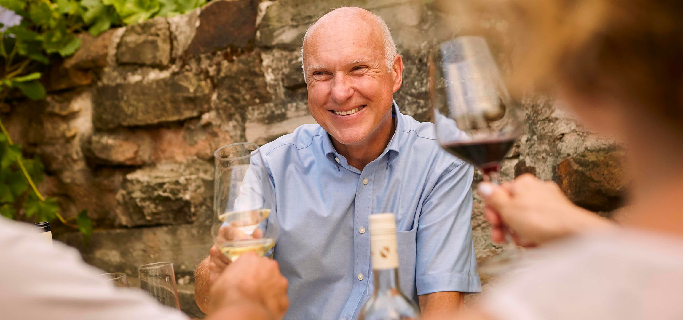 Homme souriant dégustant du vin rouge en extérieur avec un mur de pierre et de la verdure.