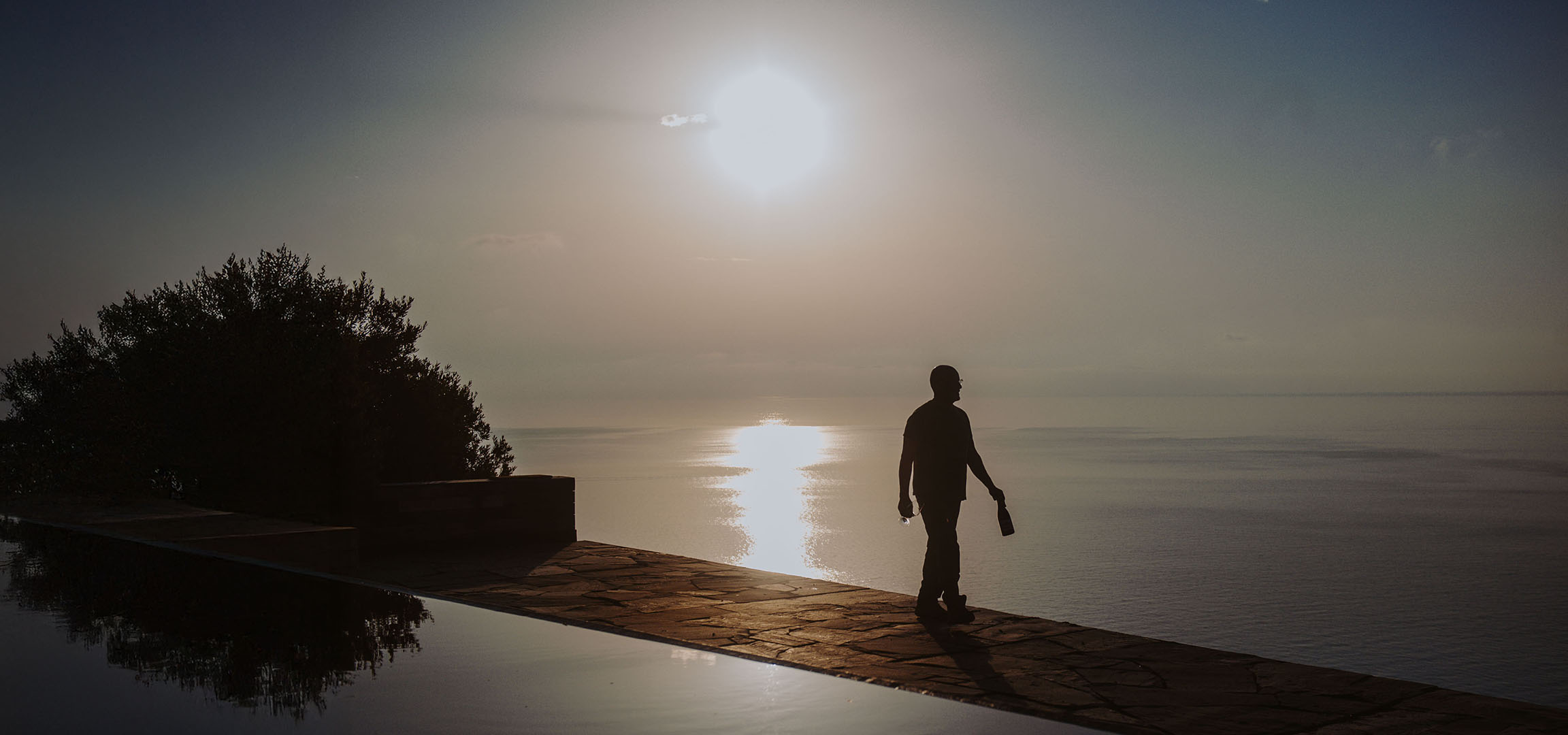 Silhouette d'un homme avec une bouteille et un verre au bord d'une piscine avec vue sur mer au coucher du soleil.