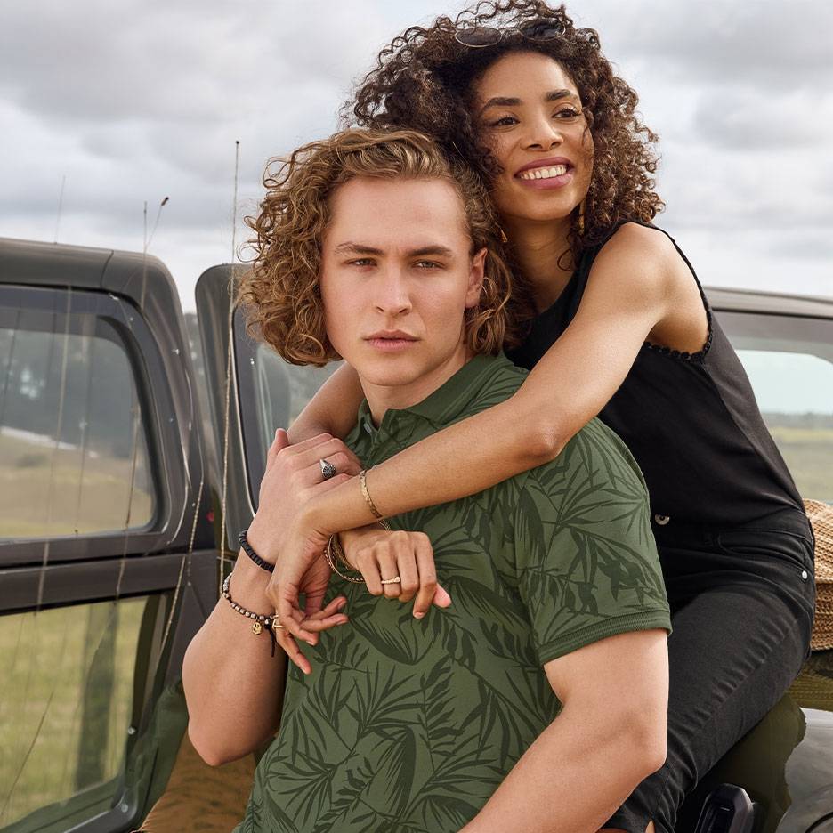 Couple souriant, lui en polo vert à motifs de feuilles, elle en débardeur noir, près d'une voiture.
