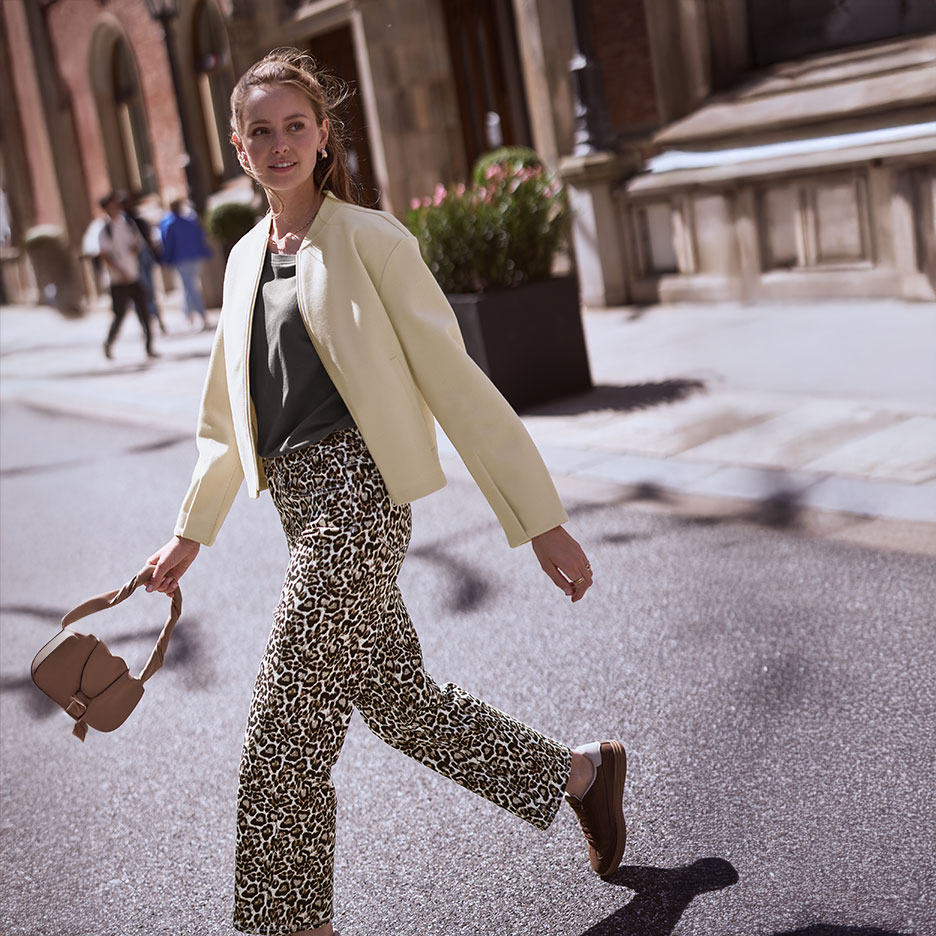 Femme en veste beige, t-shirt vert et pantalon léopard, marchant dans la rue avec un sac à main.