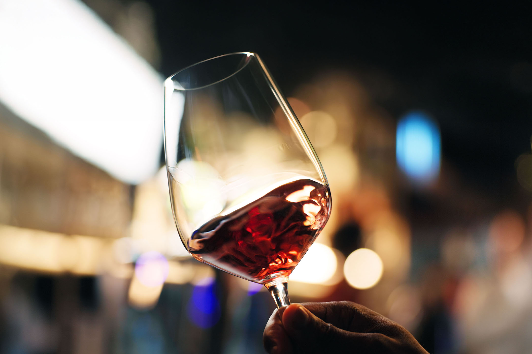 Verre de vin rouge tenu à la main, avec des lumières floues en arrière-plan.