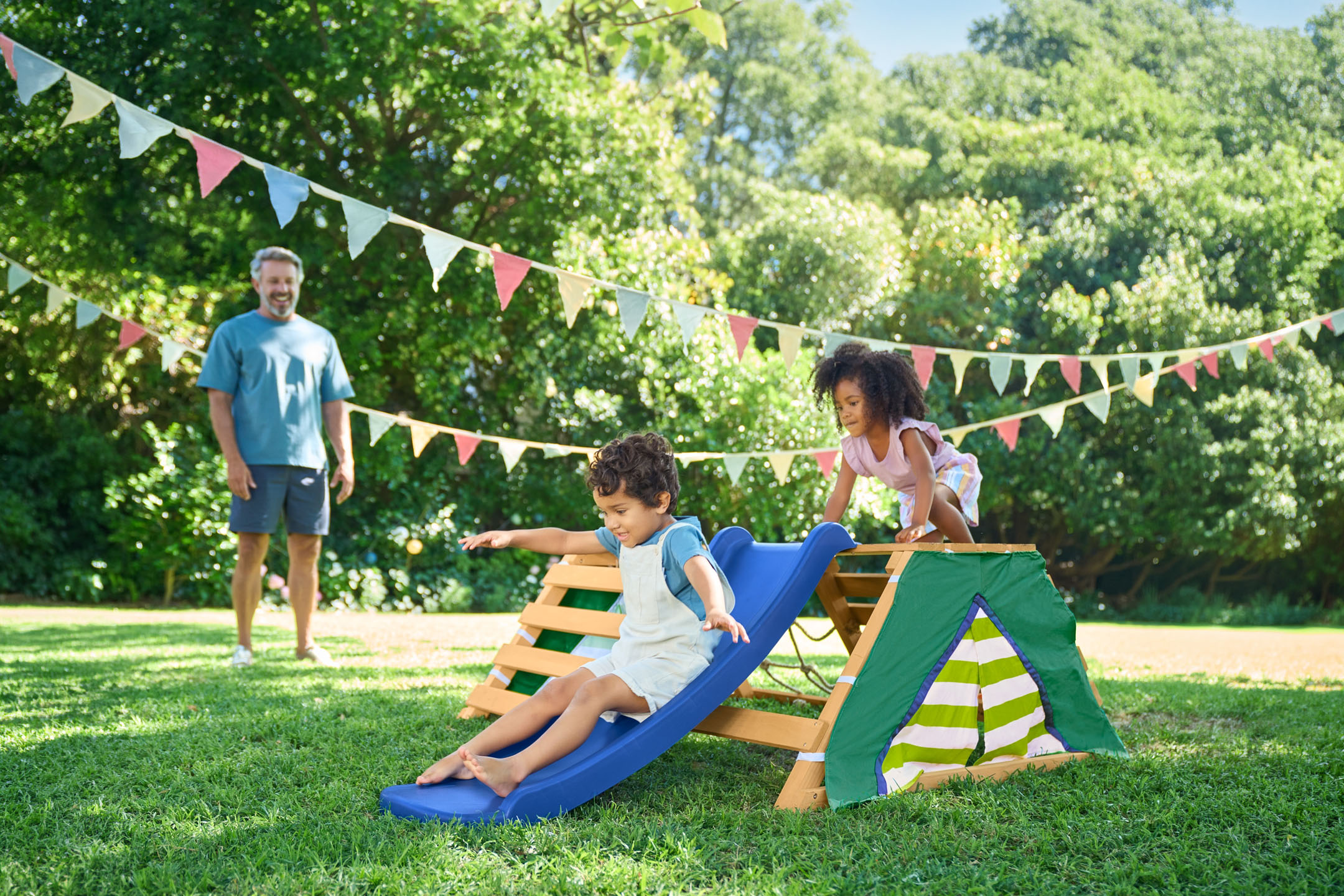 Enfants jouant sur une aire de jeux en bois avec toboggan dans le jardin, avec leur père en arrière-plan.