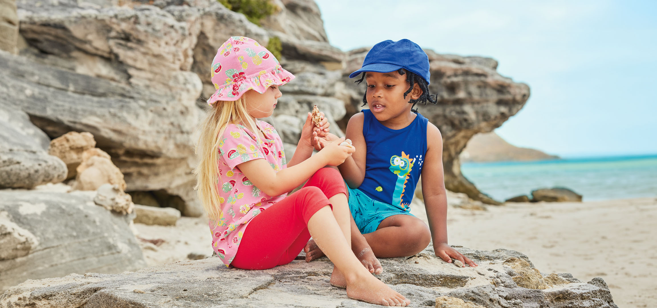 Deux enfants en tenue d'été sont assis sur un rocher près de la plage, tenant un coquillage.