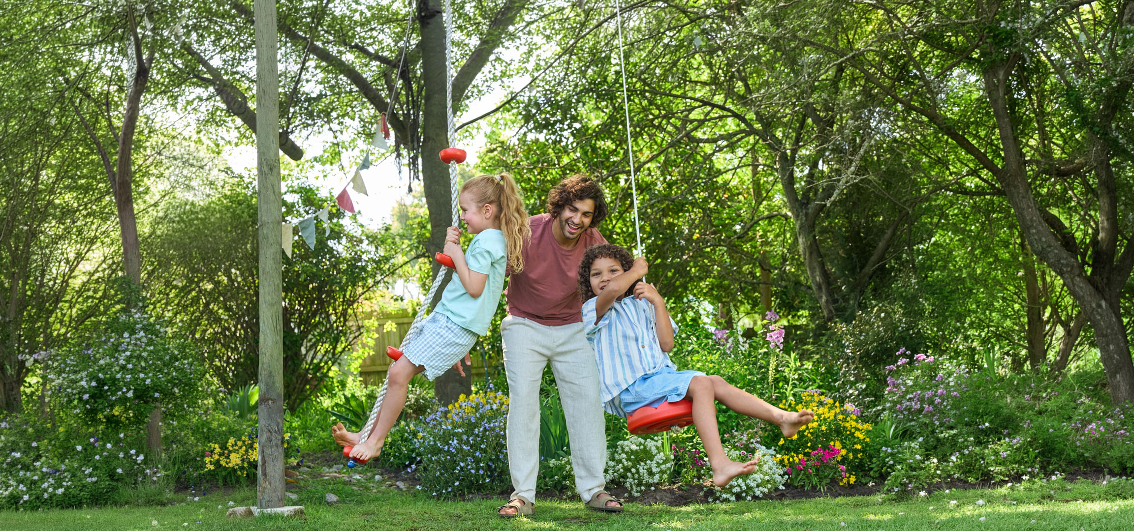 Père et deux enfants jouant sur des balançoires dans un jardin verdoyant.