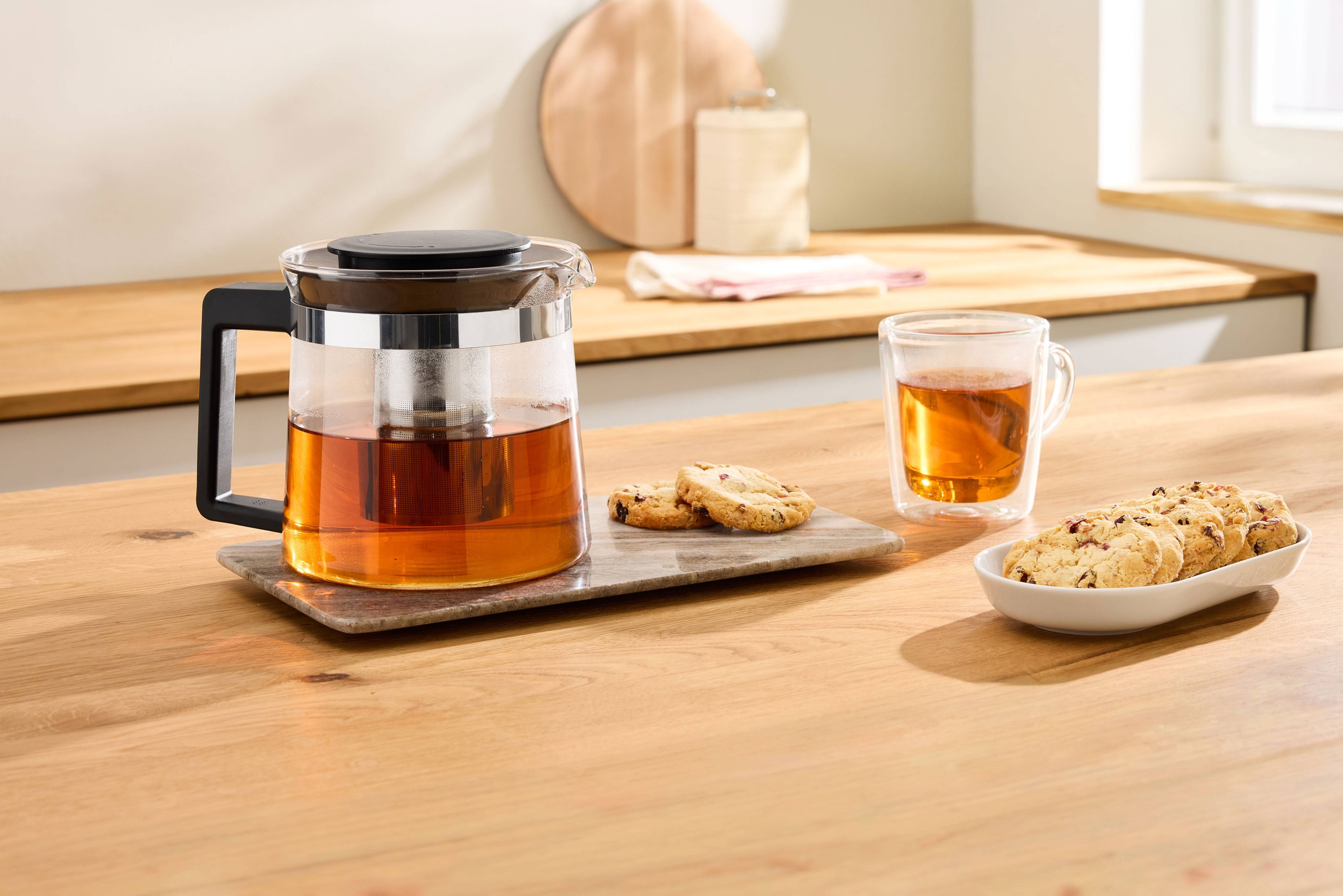 Théière en verre avec du thé et des biscuits sur un comptoir de cuisine en bois.