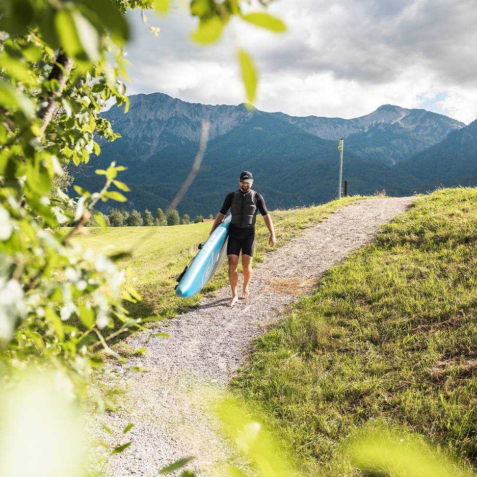 Homme en combinaison et gilet de sauvetage portant un paddleboard dans un paysage montagneux.