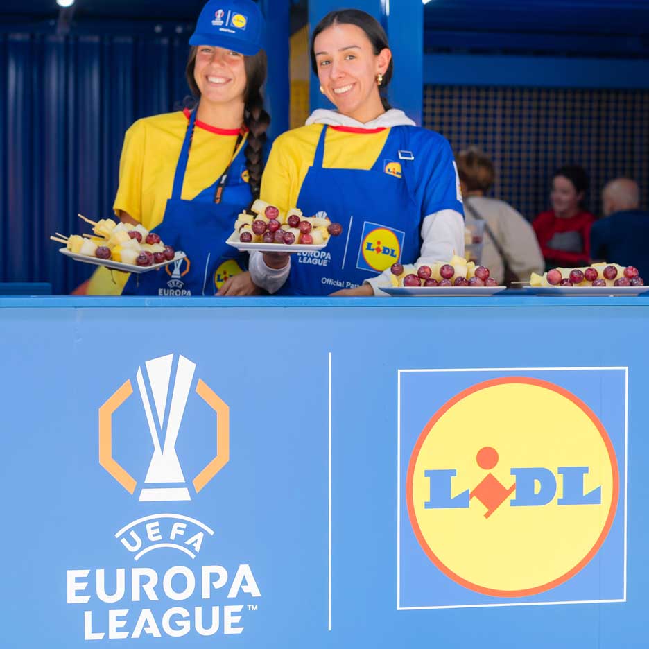 Deux femmes souriantes en tabliers Lidl offrent des brochettes de fruits lors d'un événement UEFA Europa League.