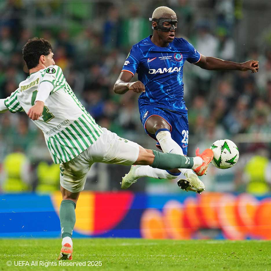 Deux footballeurs se disputent le ballon sur le terrain, l'un en blanc et vert, l'autre en bleu avec un masque.