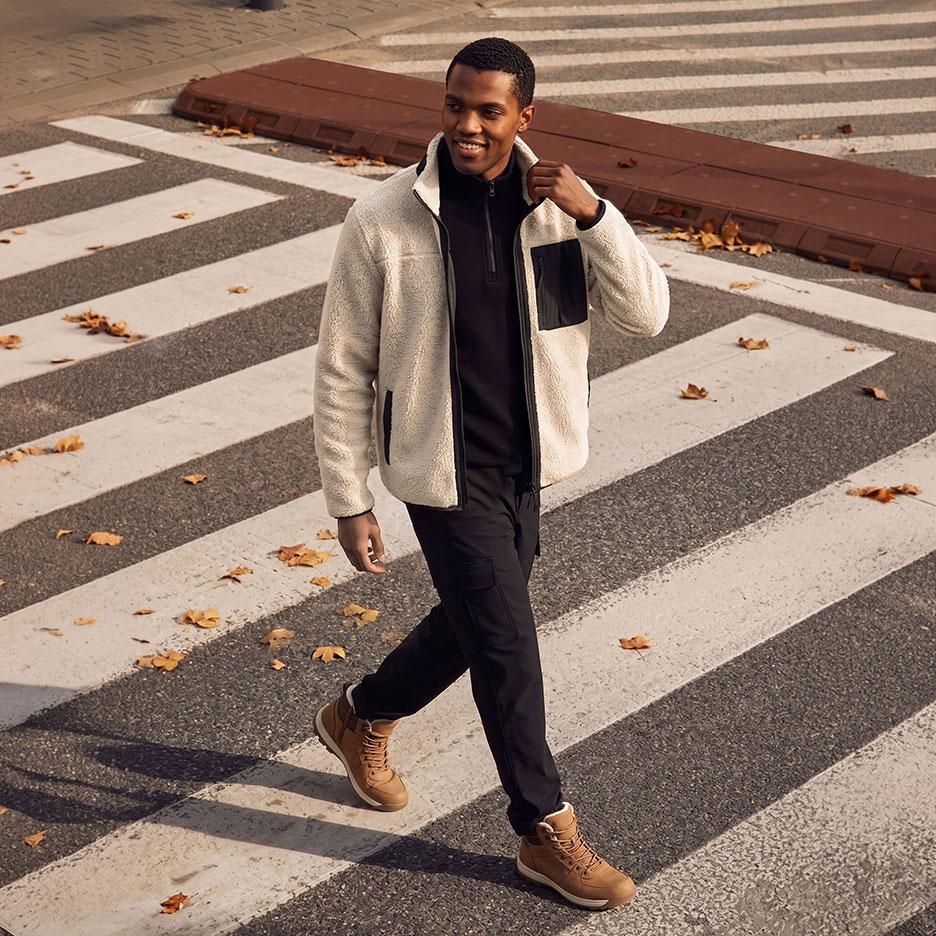 Homme souriant en veste polaire claire, pull noir et pantalon cargo noir, marchant sur un passage piéton.