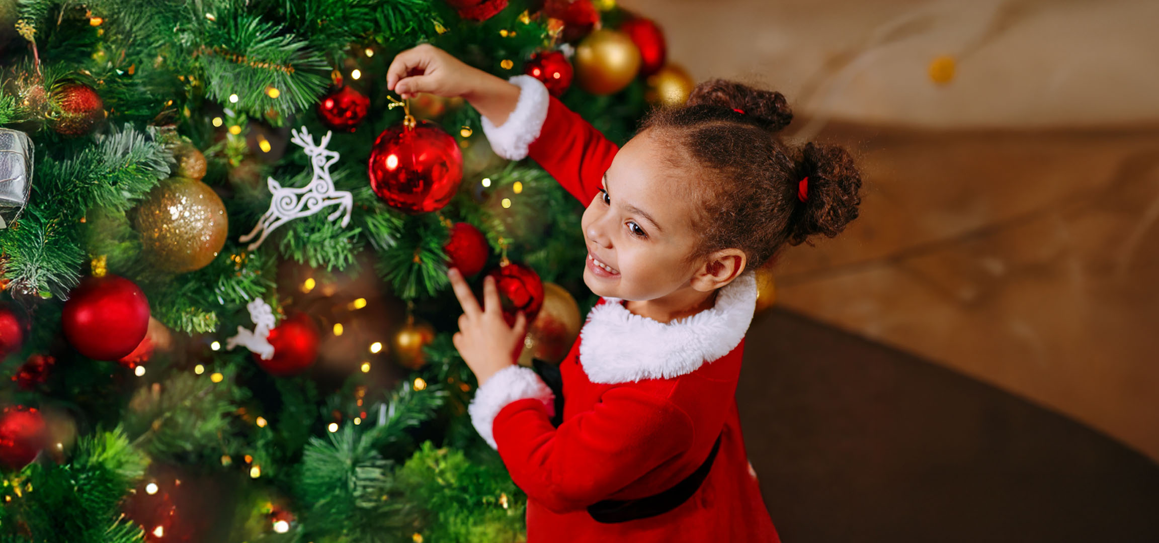 Joyeuse petite fille en costume de Père Noël décorant un sapin avec des boules rouges.