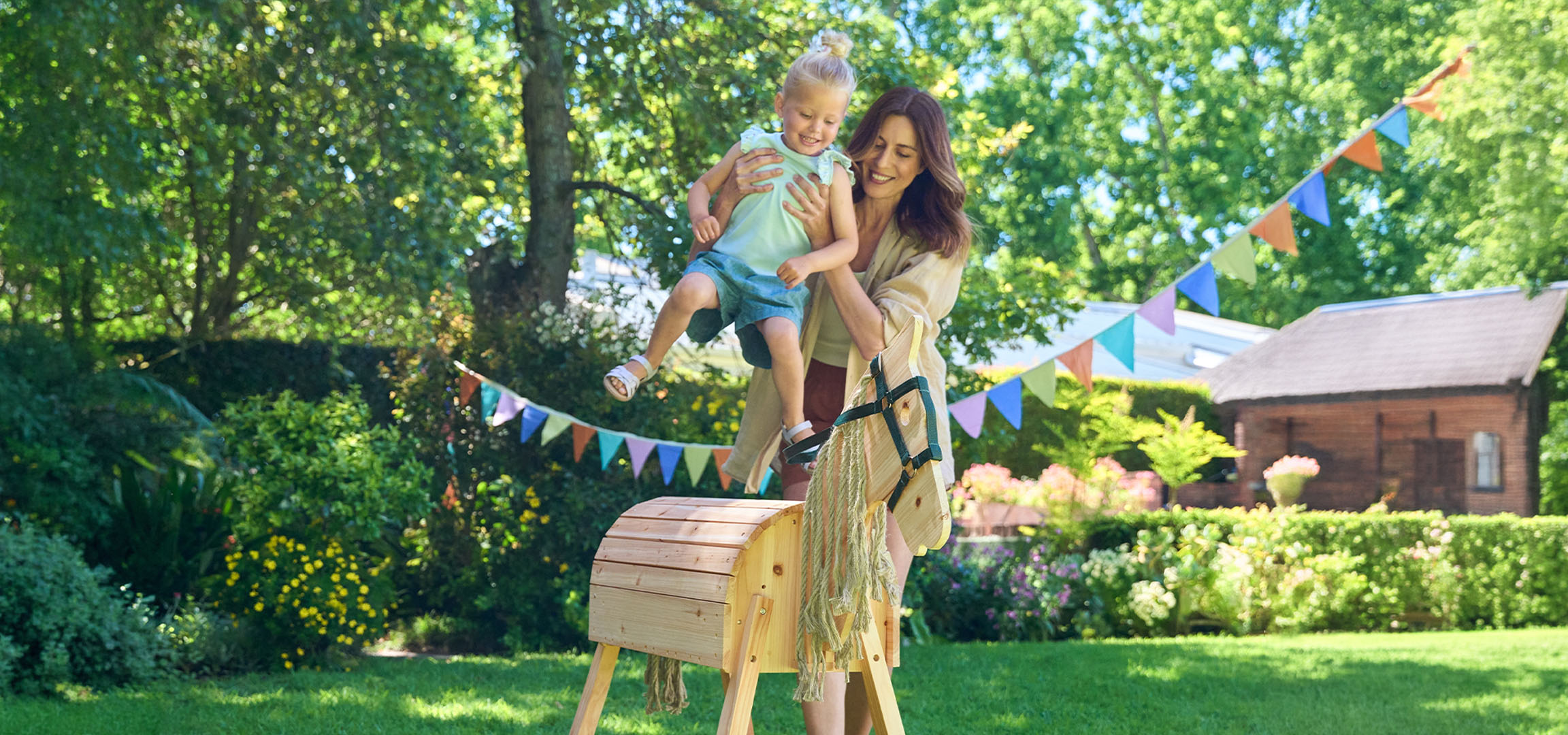 Mère et enfant jouent avec un cheval à bascule en bois dans un jardin ensoleillé.