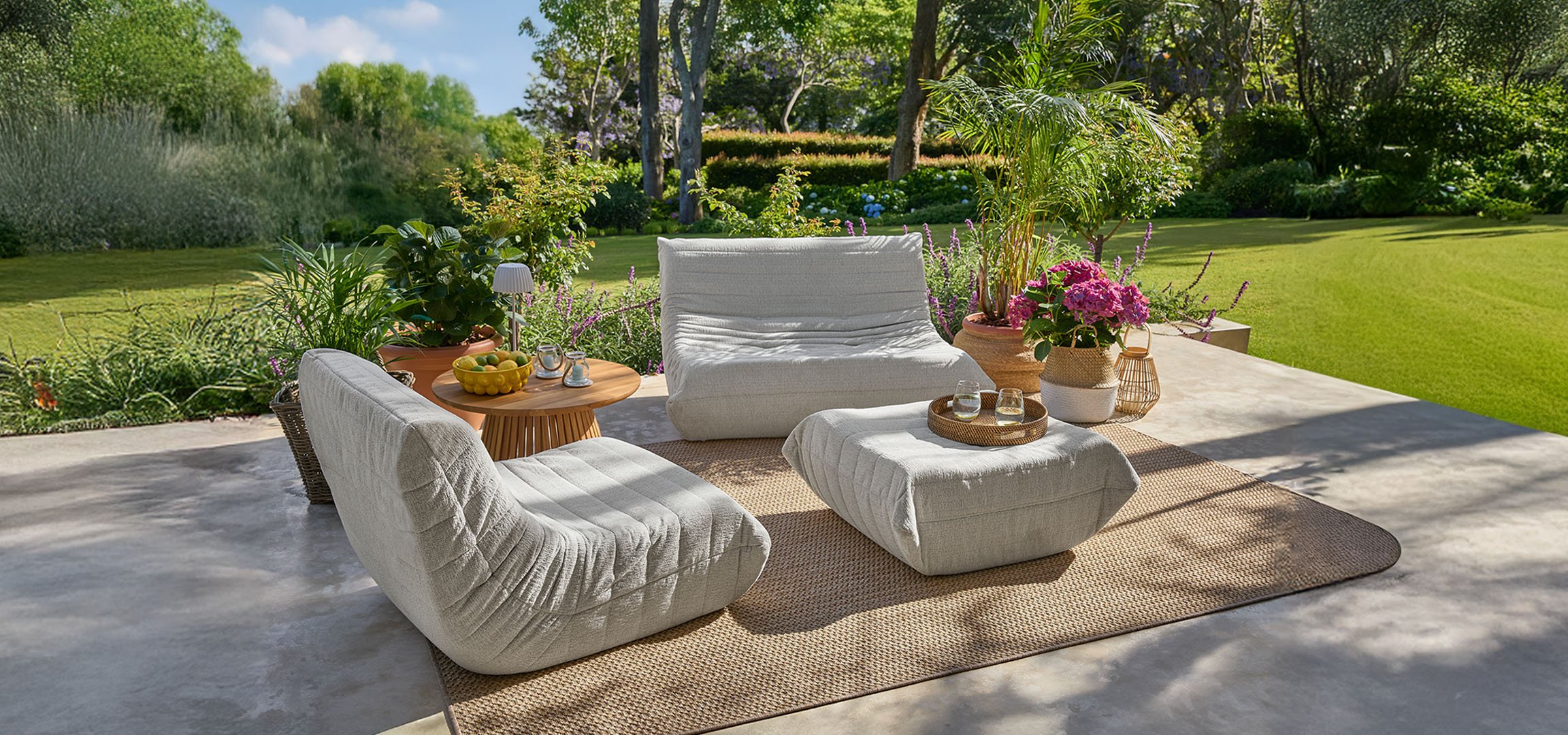 Fauteuils et poufs gris clair sur une terrasse avec une table en bois et pelouse verte.