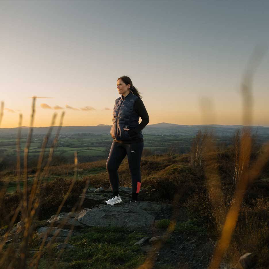 Femme en gilet matelassé et leggings debout sur une colline au coucher du soleil.