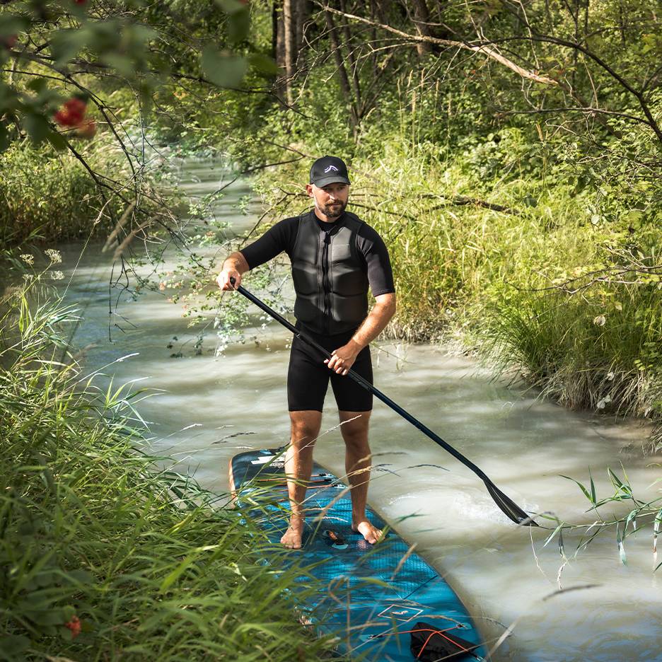 Homme en combinaison et gilet de sauvetage debout sur un paddleboard dans un ruisseau.