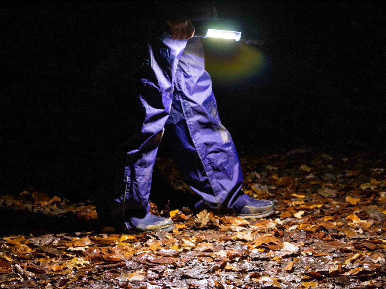 Personne en pantalon imperméable bleu marchant la nuit, éclairant les feuilles mortes avec une lampe de poche.