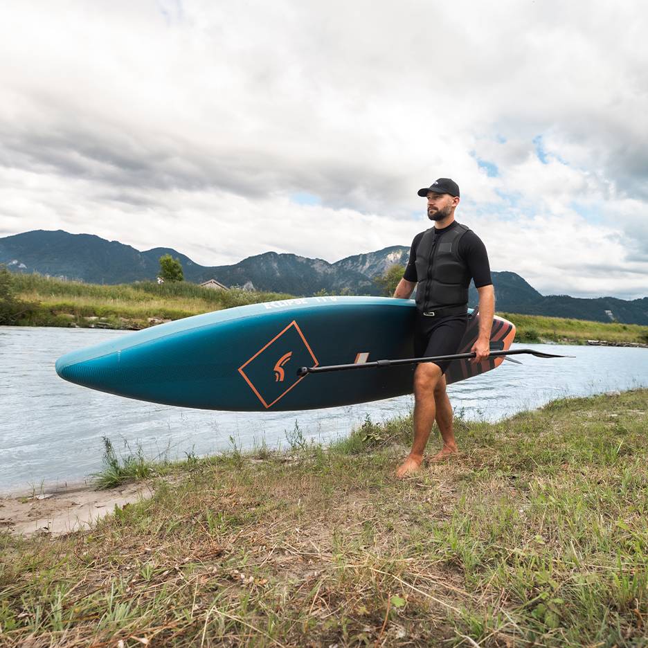 Homme en gilet de sauvetage et casquette, portant un paddleboard et une pagaie au bord d'une rivière.