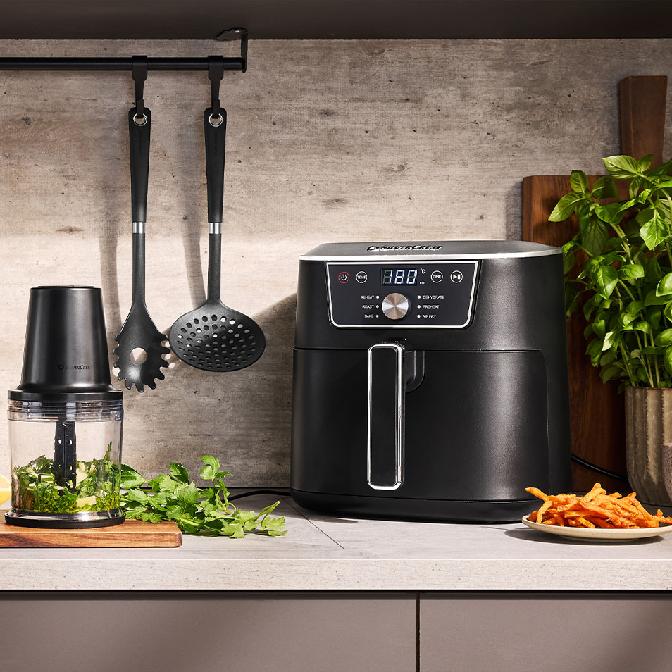 Friteuse à air SilverCrest et hachoir sur un comptoir de cuisine avec des herbes fraîches et des frites de patate douce.