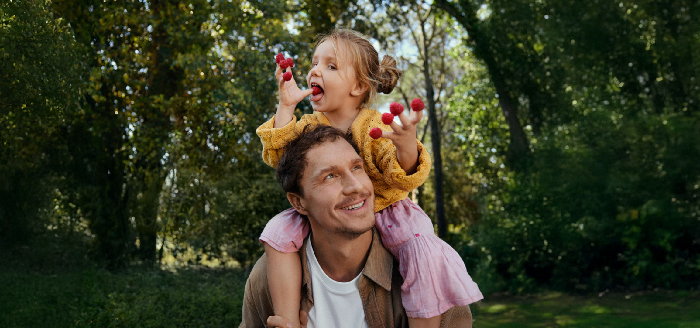 Un père souriant porte une petite fille sur ses épaules, qui mange des framboises dans une forêt.