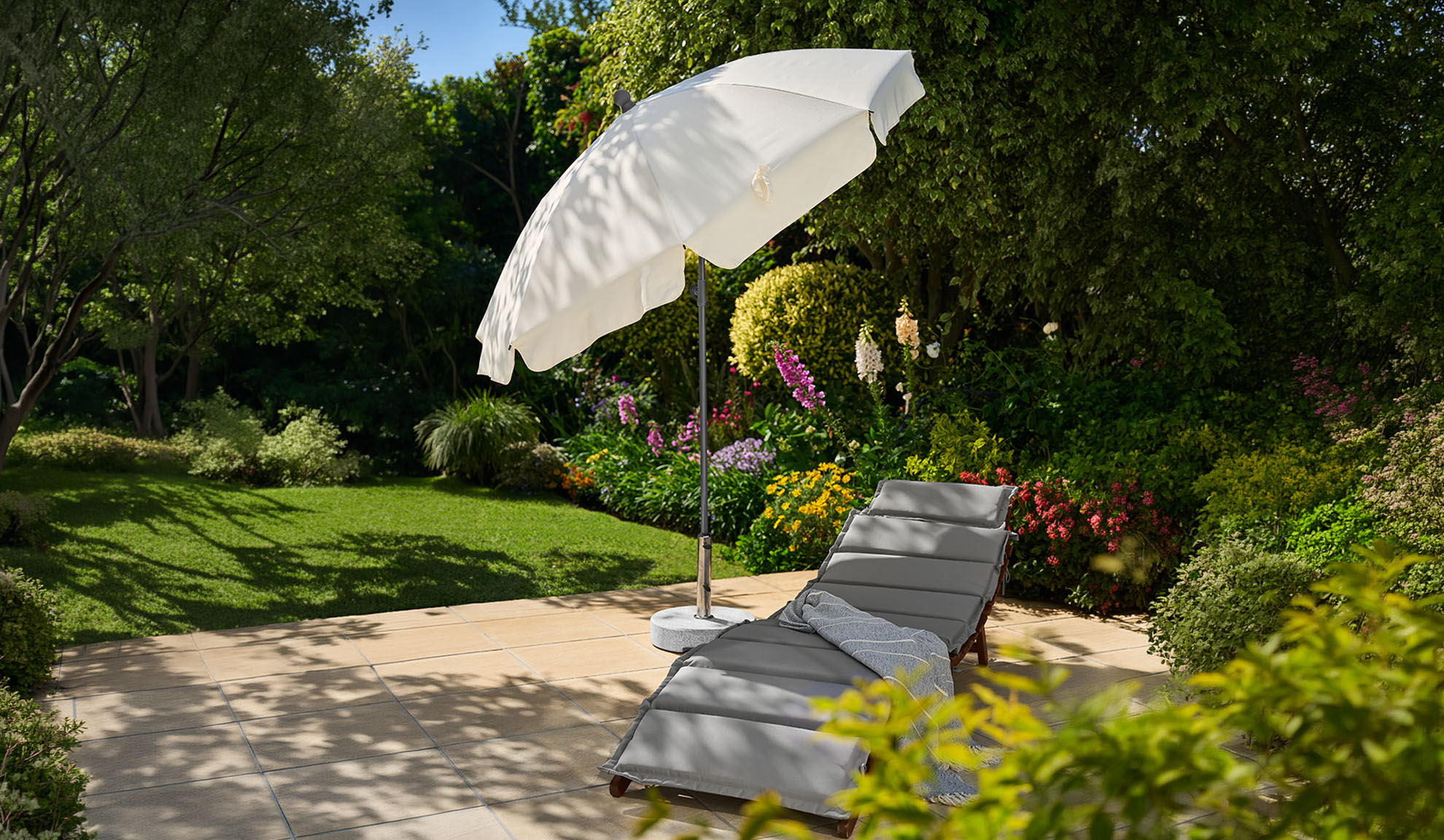 Transat gris avec parasol blanc sur une terrasse ensoleillée avec jardin luxuriant.