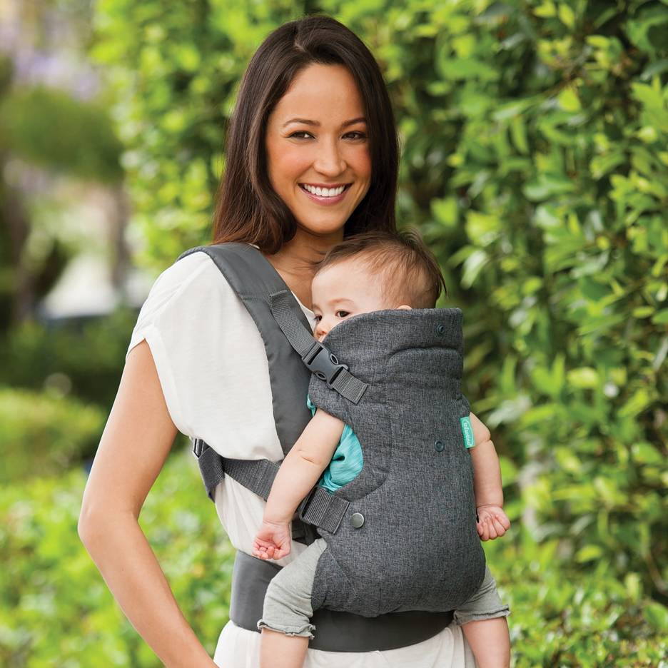 Femme souriante dans un porte-bébé gris tenant un bébé en extérieur.