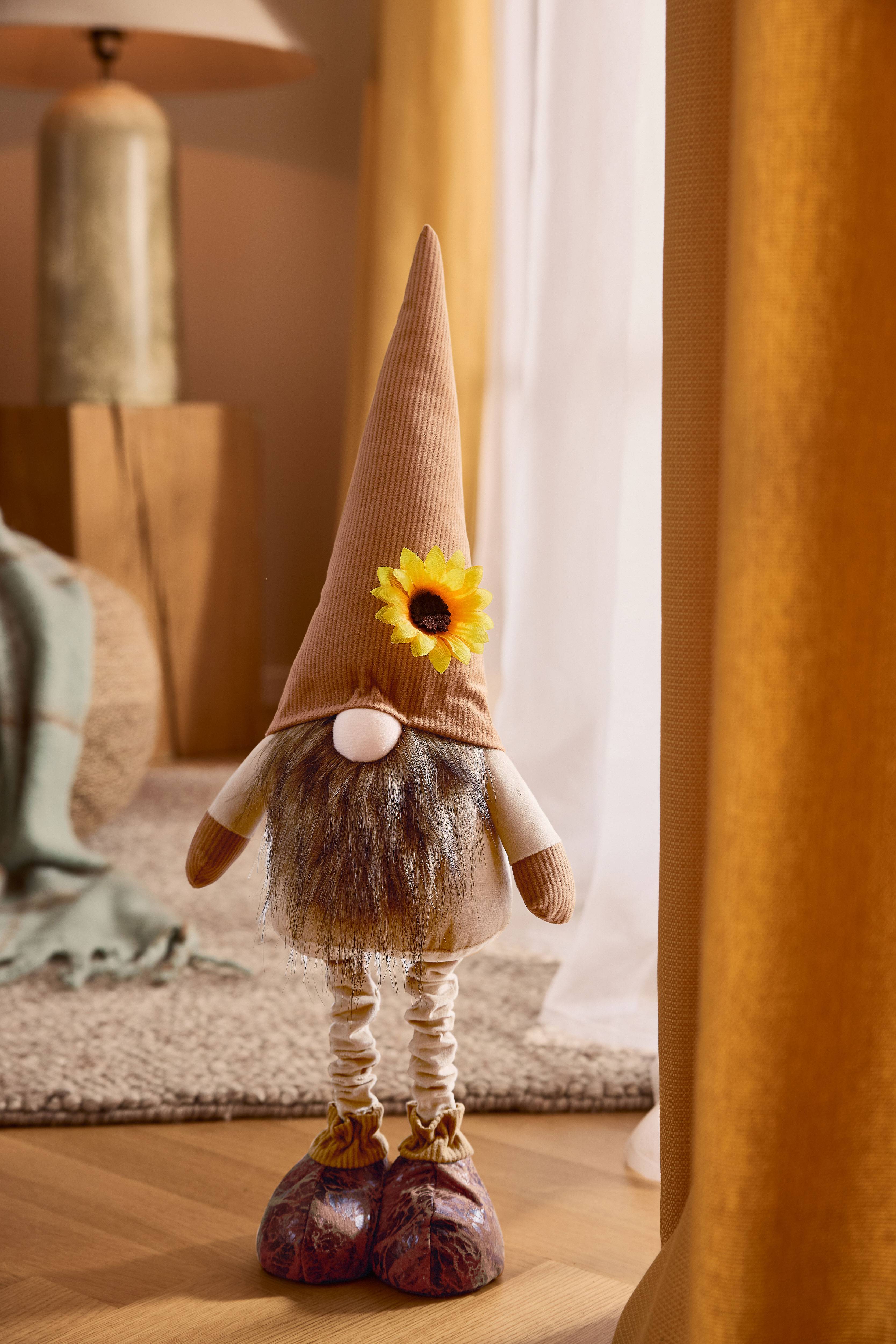 Gnome en peluche avec un chapeau pointu marron, une barbe et un tournesol, dans un cadre douillet.