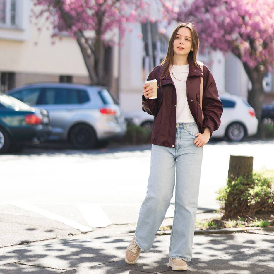 Femme en veste et jean avec café dans une rue aux arbres en fleurs.
